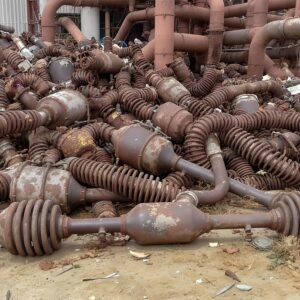 Rusted car parts including mufflers, springs, and axles dumped on concrete ground, with industrial pipes in background