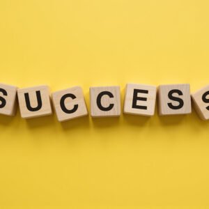 Wooden letter cubes spelling SUCCESS arranged in a loose uneven row each cube slightly tilted at a different angle bold black letters bright yellow background flat lay minimal business success concept